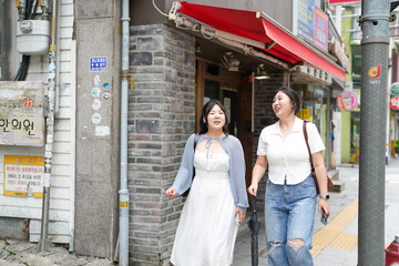Two Korean women in their twenties in casual clothes walk along a boulevard sidewalk, talking and...