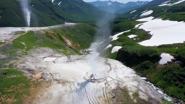 Aerial view of geysers erupting steam in a valley surrounded by green mountains with patches of snow, Kamchatka Krai, Russia.