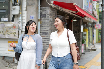 Two Korean women in their twenties in casual clothes walk along a boulevard sidewalk, talking and...