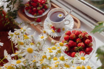Freshly picked strawberries are displayed in elegant bowls alongside a warm cup of tea. The sunny windowsill, adorned with vibrant daisies, creates a cozy and inviting atmosphere for relaxation