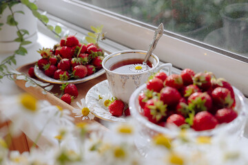 Delightful arrangement featuring ripe strawberries in bowls alongside a delicate cup of tea, all set against a bright and cheerful window backdrop filled with blooming flowers