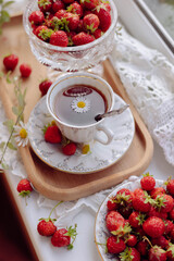 A wooden tray is adorned with two bowls of red strawberries alongside a delicate cup of tea topped with a daisy. The setting evokes a warm and inviting afternoon gathering at home