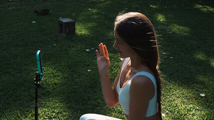 Yoga instructor waving and greeting students into phone camera for online class in city park while...