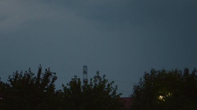 A thunderstorm brews with dark clouds and occasional lightning illuminating the blue sky. Nearby trees sway in the wind as rain begins to fall, signaling severe weather.