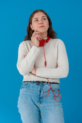 Pensive young woman with red headphones thinking and looking up, standing against a solid blue...