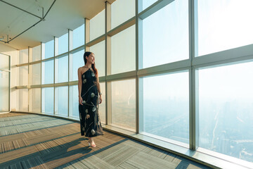 Fototapeta premium Beautiful asian stylish woman in elegant dress standing in front of glass window inside modern highrise building with city skyline at Bangkok