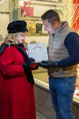 Couple admiring a Merry Christmas gift box near a decorated store window.