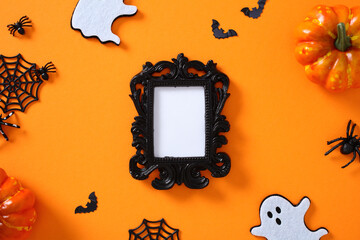 Halloween flat lay composition on bright orange background with ornate black frame, pumpkins, ghost cutouts, spiders, webs, and bats