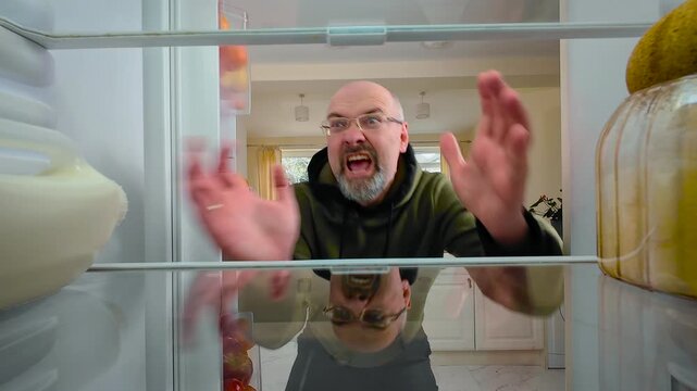 Man opening fridge and screaming at emptiness with furious expression, view from inside of fridge. Male reacting to hunger and lack of food while feeling enraged and desperate. Guy yelling in