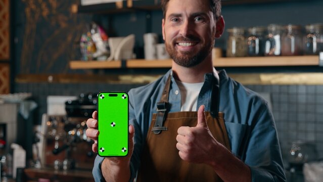 Satisfied happy Caucasian barista man showing green screen mobile phone thumb up in cafeteria cafe male small business entrepreneur recommend application advertisement guy waiter approval catering app - Powered by Adobe