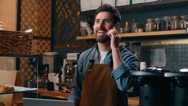 Happy Caucasian guy employee talking mobile phone male man barista using computer speak with client delivery small business owner entrepreneur cheerful waiter work in cafeteria restaurant multitasking