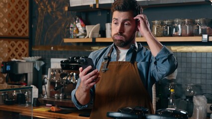 Frustrated confused Caucasian barista man look screen mobile phone in cafeteria male small business owner upset bartender using smartphone waiter guy money loss difficulty online trouble failure cafe