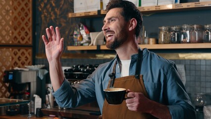 Cheerful happy Caucasian waiter cafe worker barista small business owner holding cup of coffee beverage drink male smile in coffeehouse friendly man in apron greeting client guy waving hand catering