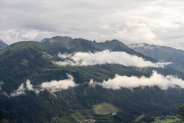 Majestic alpine peaks with snow caps, forested green slopes, and clouds floating under dramatic summer sky
