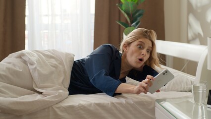 Sleepy Caucasian woman wake up in bed check time mobile phone overdue in bedroom morning shocked...