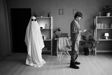 Man looks at old photo of deceased wife in quiet room, unaware of female ghost standing beside him....