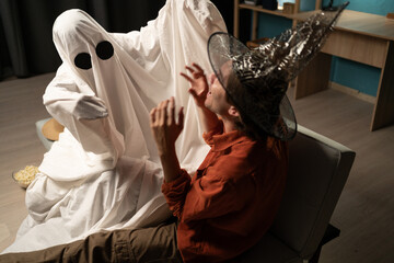 Man in shirt and ghost-costumed person having fun together sitting on sofa at home during Halloween...