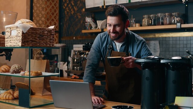 Glad Caucasian entrepreneur staff work in cafe waiter smile drink cup coffee energy beverage man typing laptop restaurant guy male barista bartender employee ordering supplies Internet small business