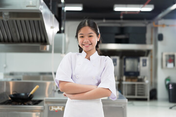 Restaurant kitchen. Asian child girl chef standing with crossed arms in kitchen, wearing white...