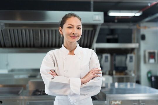 Restaurant kitchen. Female chef standing with crossed arms in kitchen, wearing white apron and smiling looking to camera
