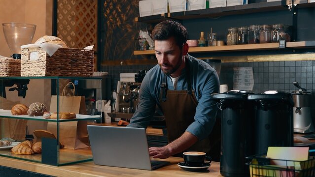 Glad Caucasian entrepreneur staff work in cafe waiter smile drink cup coffee energy beverage man typing laptop restaurant guy male barista bartender employee ordering supplies Internet small business - Powered by Adobe