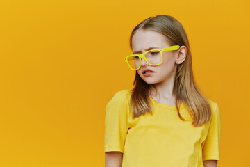 A young girl with long hair wearing yellow glasses and a yellow t shirt against a bright yellow...