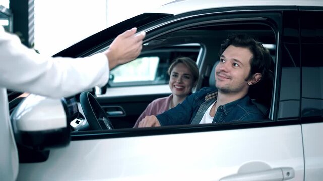 Happy couple at car dealership receiving keys from salesperson, celebrating successful purchase of new vehicle with excitement and satisfaction