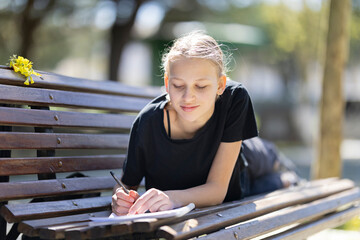 Teenager girl doing her homework lying on a bench in a park