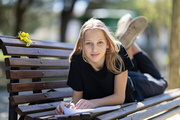 Young student girl lying on bench writing in notebook in park