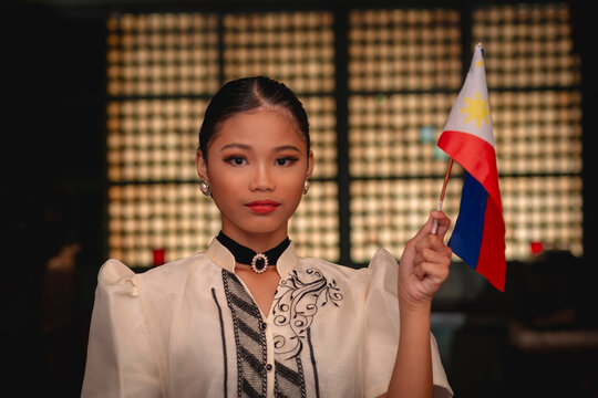 Young Filipina woman in traditional attire confidently holds Philippine flag inside an old heritage home with capiz windows.