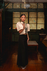 Graceful Filipina woman in Filipiniana poses with hand on chest inside antique Filipino home with wooden floors and capiz panels.