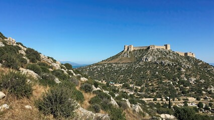 Fototapeta premium Landscape showing a steep rocky incline covered with shrubs and grass leading to an ancient castle with defensive walls and towers on a mountain summit under a bright blue sky