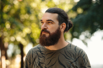 A man with a beard and long hair stands outdoors, deeply lost in thought and reflection, amidst...