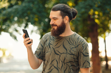 A bearded man checks his phone outdoors in a serene, green environment, showcasing modern lifestyle.