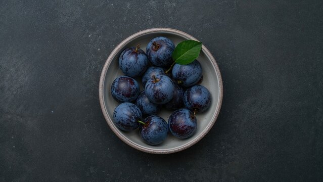 Flat lay of blue plums from the garden in a bowl set on a dark concrete backdrop - Powered by Adobe