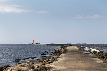 View of a long concrete pier in the sea with a lighthouse at the end. Large cargo ships are visible in the distance on the horizon