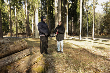 Two people dressed in winter jackets standing in the woods, pausing for a warm drink from a thermos, enjoying nature, quiet conversation, and seasonal coziness.