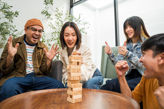 Young Asian friends are happily playing wooden block together at home, enjoying their leisure time. Group friends carefully remove a wooden block while others watch, having fun a classic table game.