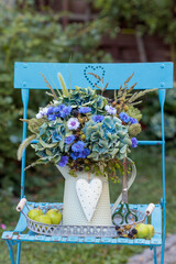 Blumenstrauß mit blauen Hortensienblüten, Federnelken, Wilder Möhre, Gräsern und Brombeerzweigen im vintage Krug auf einem Gartenstuhl