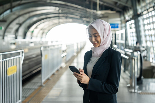 Muslim asian woman using smartphone while waiting on platform at train station in the city. Beautiful islamic female checking application on cell phone - Powered by Adobe