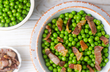 Boiled green peas with fried crispy bacon in a white ceramic bowl on the table. Concept of an old recipe. Simple dish. Horizontal orientation. Selective focus. Top view.