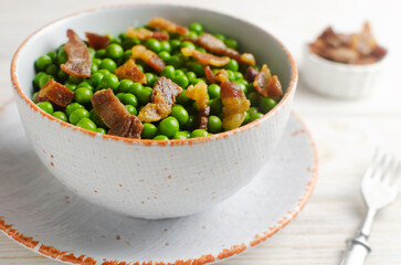 Boiled green peas with fried crispy bacon in a white ceramic bowl on the table. Concept of an old recipe. Simple dish. Horizontal orientation. Selective focus.