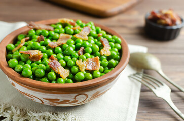Boiled green peas with fried crispy bacon in a brown ceramic bowl with a fork and spoon on a wooden table. Old recipe concept. Simple dishes. Rustic style. Horizontal orientation. Selective focus.