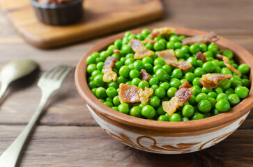 Boiled green peas with fried crispy bacon in a brown ceramic bowl with a fork and spoon on a wooden table. Old recipe concept. Simple dishes. Rustic style. Horizontal orientation. Selective focus.