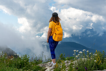 Young woman with a yellow backpack stands on a rocky ledge, gazing at a breathtaking mountain...