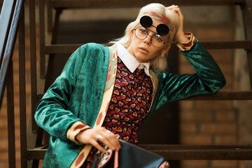 A fashionable woman with striking glasses poses on a staircase, showcasing her unique style.