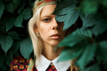 A young woman with blonde hair poses amongst lush green leaves.