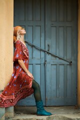 A woman in a flowing floral dress leans by a weathered blue door, creating a dreamy and stylish...