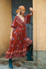 Bohemian woman in a stylish floral dress and boots leans against a wall, outdoor.