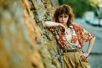A stylish young woman with curly hair poses by a stone wall, looking at the camera.